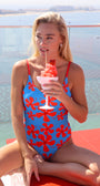 Woman in a blue and red swimsuit holding a watermelon smoothie by a pool.
