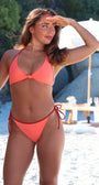 Woman in a coral bikini standing on a beach with an umbrella and table in the background.