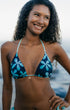 A woman with curly dark hair smiling outdoors, wearing a navy bikini top with a light blue floral pattern and a small gold necklace.