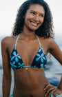 A woman with curly dark hair smiling outdoors, wearing a navy bikini top with a light blue floral pattern and a small gold necklace.