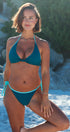 A woman posing on a sandy beach wearing a two-piece bikini. The bikini features a deep teal blue top and bottom with a contrasting light aqua blue trim and side ties.