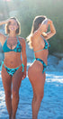 Two women modeling matching bikini swimsuits on a sandy beach. The swimsuits feature a teal and turquoise floral pattern, with one model showing the front view and the other showing the side/back view.