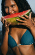 Woman in teal reversible bikini eating a watermelon by the ocean