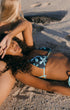 Two women on a sandy beach wearing matching blue and white floral print string bikinis, with one woman lying down and the other sitting behind her in warm sunlight.