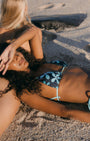 Two women on a sandy beach wearing matching blue and white floral print string bikinis, with one woman lying down and the other sitting behind her in warm sunlight.