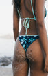 Rear view of a woman wearing a bikini on a beach, with sand on her skin. The bikini has a dark blue base with a light blue and white floral pattern on the bottom and light blue straps.